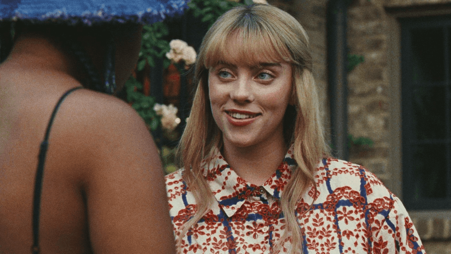 Billie Eilish smiles in her blonde hair and flower print shirt as she talks with Dominique Fishback in 'Swarm.'